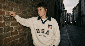 Person wearing a 'USA Soccer' sweater leaning against a brick wall in an urban setting.